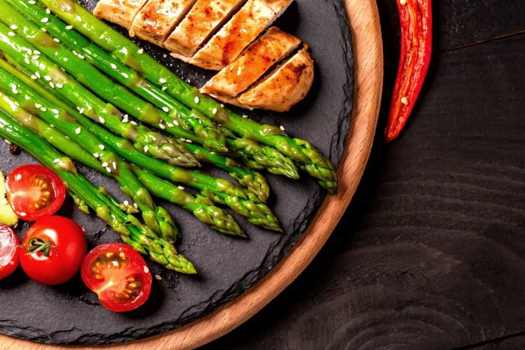 פרוסות עוף בגריל, אספרגוס ירוק עז ועגבניות שרי חצויות עם שומשום מסודרות יפה על צלחת צפחה עגולה עם שוליים עץ. פלפל צ'ילי אדום מונח ליד הצלחת על משטח עץ כהה.