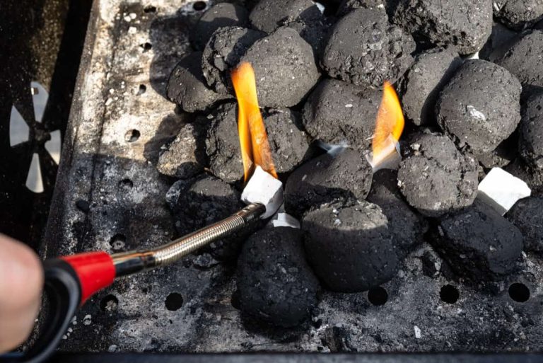 יד אוחזת מצית מדליקה קובייה לבנה מתחת לבריקט פחם שחור בגריל, עם להבות קטנות גלויות. חתיכות הפחם מוערמות ובעיקר לא מוארות.