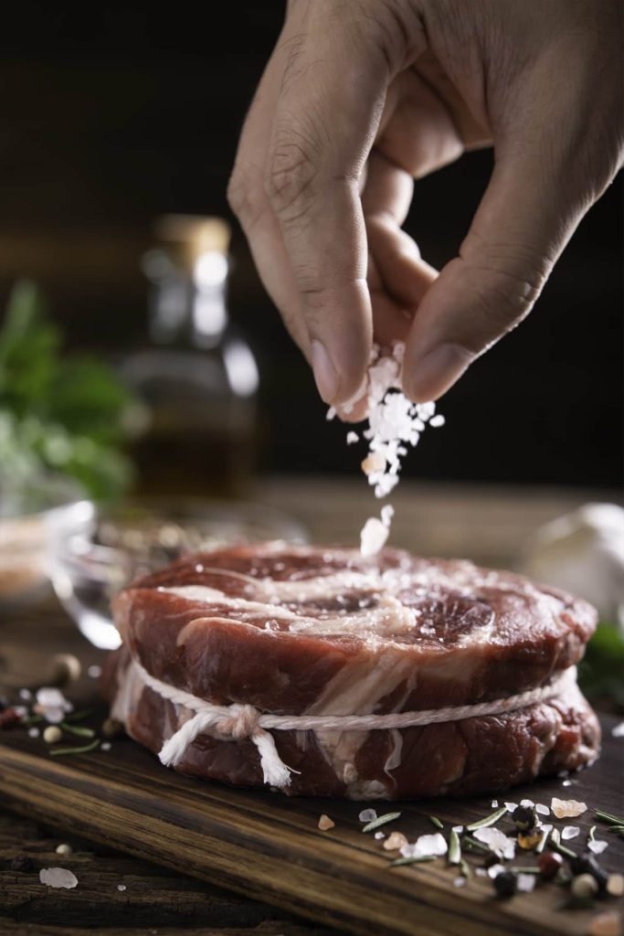 יד מפזרת מלח על שני סטייקים נאים קשורים בחוט על לוח עץ. עשבי תיבול וגרגרי פלפל מפוזרים מסביב. בקבוק וירוקים מטושטשים ברקע.