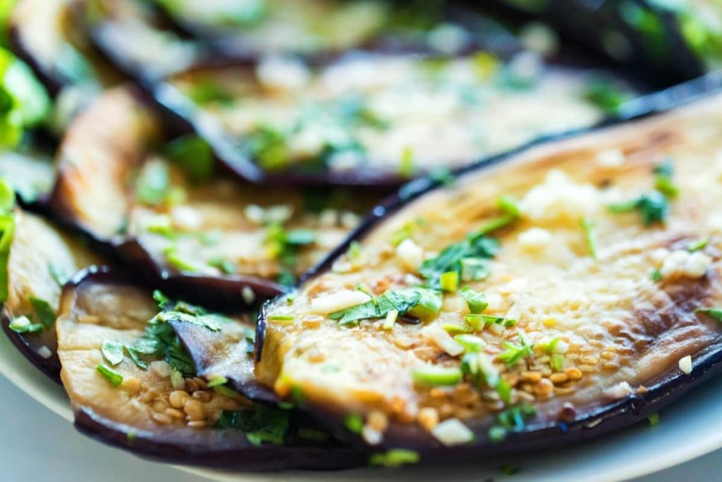 תקריב של חציל בגריל פרוס מעוטר בעשבי תיבול קצוצים ושום על צלחת לבנה. החציל חום זהוב עם מעט פחם, והירוקים מוסיפים נופך רענן.