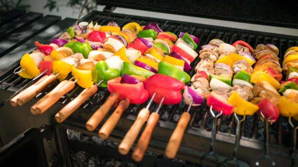 שיפודי ירקות צבעוניים עם פלפלים, פטריות, בצל ועגבניות צולים על מנגל חיצוני. לשיפודים יש ידיות עץ והם מסודרים בצורה מסודרת על רשתות הגריל.