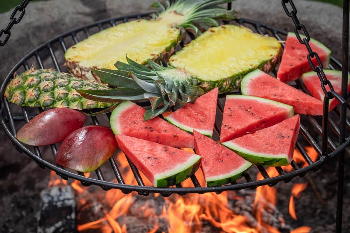 אבטיח פרוס, אננס ומנגו מסדרים על גריל עגול על להבה גלויה. הפירות הבהירים מנוגדים לגריל הכהה והאש המרצדת מתחת.