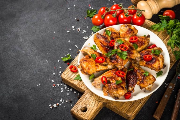 כנפי עוף בגריל בעיטור פרוסות צ'ילי אדום ופטרוזיליה על צלחת לבנה. הצלחת מונחת על קרש חיתוך מעץ. עגבניות שרי ומטחנת פלפלים ברקע על משטח כהה.