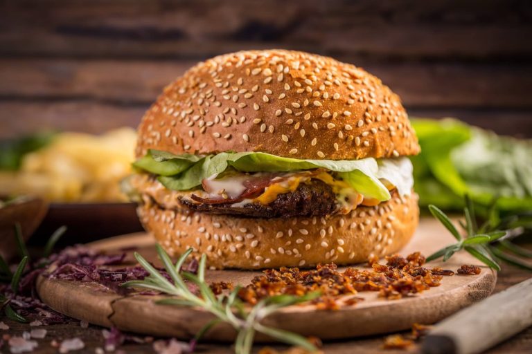 צ'יזבורגר עסיסי עם חסה, עגבנייה ורוטב יושב על לוח עץ כפרי. לחמניית השומשום בצבע חום זהוב, ומסביב פזורים חתיכות פריכות ועשבי תיבול טריים. רקע מטושטש מרמז ליותר ירוק ומרכיבים.