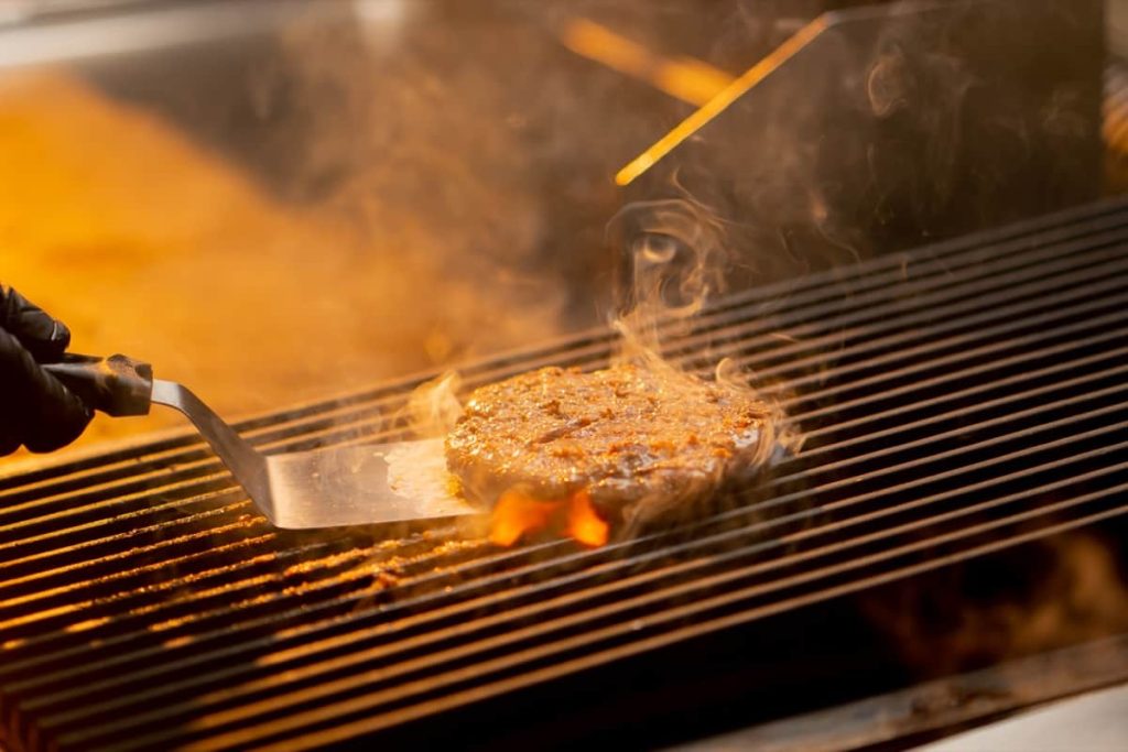 תקריב של קציצת המבורגר רוחשת על גריל עם להבות ועשן עולה. מרית המוחזקת ביד מכוסה כפפה עומדת להפוך את הקציצה. הסצנה חמימה עם זוהר זהוב מהלהבות.