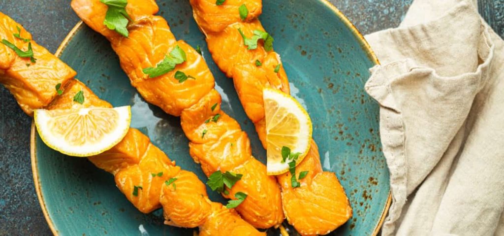 שלושה שיפודי סלמון בגריל מסודרים על צלחת טורקיז. כל שיפוד מעוטר בפטרוזיליה טרייה ופלחי לימון. מפית בד בז' נראית חלקית לצד הצלחת.