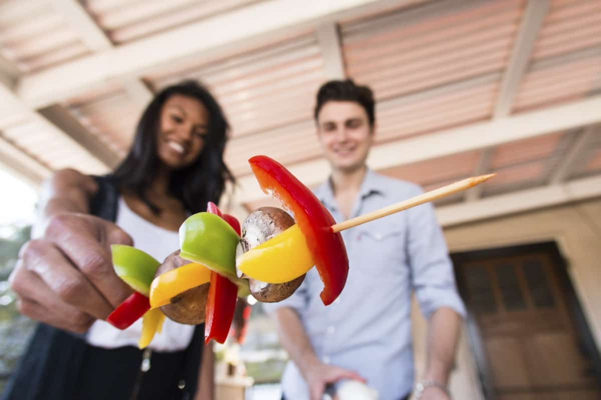 אישה וגבר עומדים מחייכים בחוץ מתחת לחופה. האישה מחזיקה שיפוד עם ירקות ופטריות צבעוניים לכיוון המצלמה. הרקע כולל בית ודלת מטושטשת.