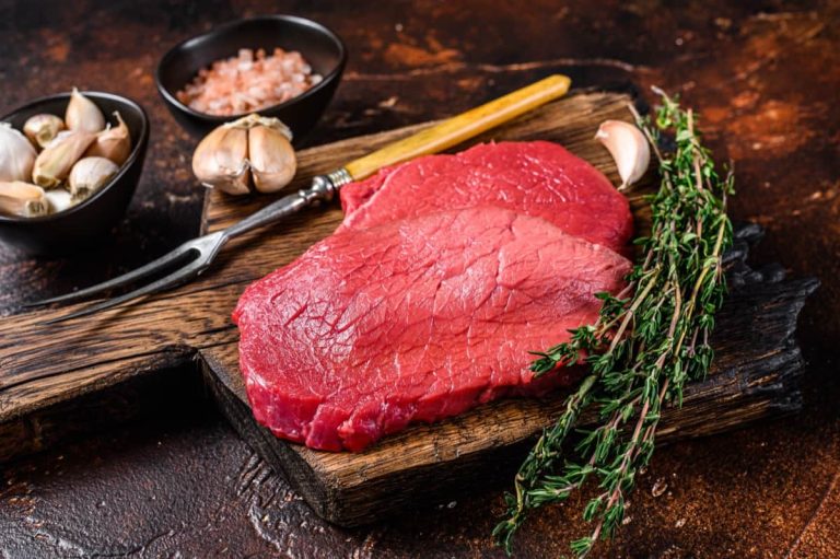 סטייקים בקר נא על קרש חיתוך עץ עם מזלג גילוף, שיני שום, קערת מלח ורוד וענפי טימין טרי על רקע כפרי כהה.