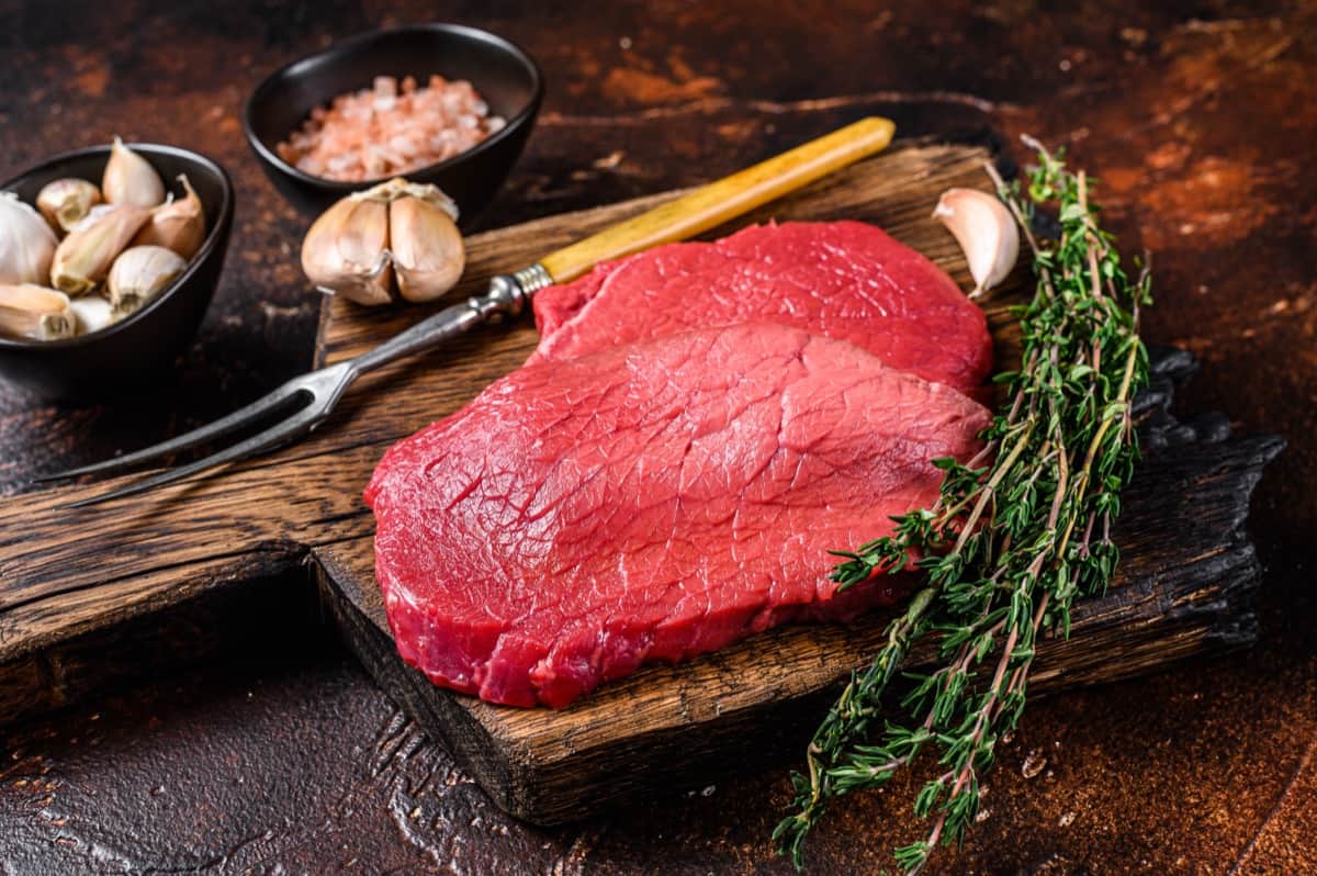 סטייקים בקר נא על קרש חיתוך עץ עם מזלג גילוף, שיני שום, קערת מלח ורוד וענפי טימין טרי על רקע כפרי כהה.