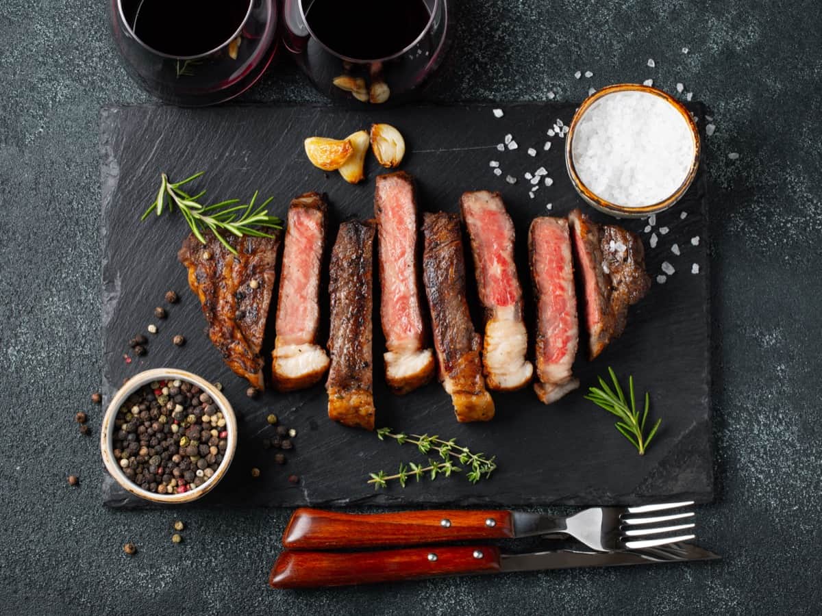 סטייק מדיום-רייר פרוס על קרש הגשה כהה עם רוזמרין, שום ומלח. קערות של גרגירי פלפל ומלח נמצאות בקרבת מקום, יחד עם שתי כוסות יין אדום. מזלג וסכין מונחים ליד הסטייק.