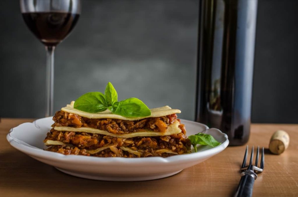 צלחת לזניה עם בזיליקום טרי מונחת על שולחן עץ. לצדו, מזלג ובקבוק יין עם פקק. כוס יין אדום נמצאת ברקע, על רקע כהה ומרקם.