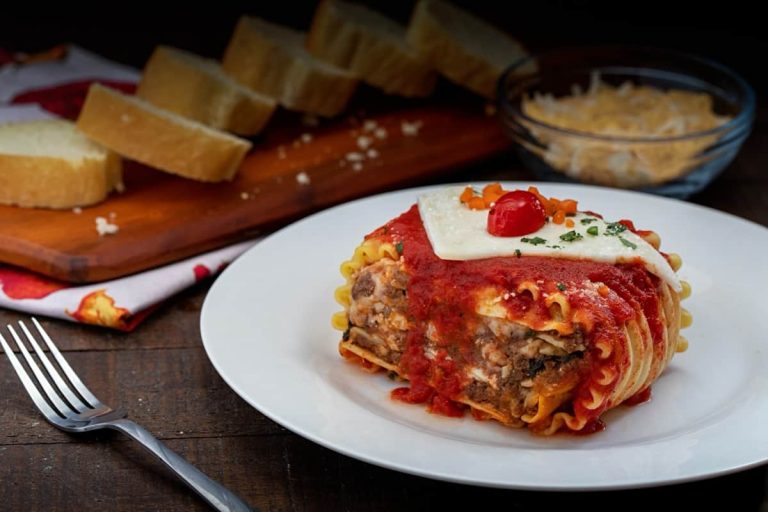 צלחת עם פרוסת לזניה בתוספת רוטב עגבניות, עגבנייה שרי וגבינה מותכת. ברקע לחם פרוס וקערת גבינה מגוררת על לוח עץ. מזלג מונח ליד הצלחת.