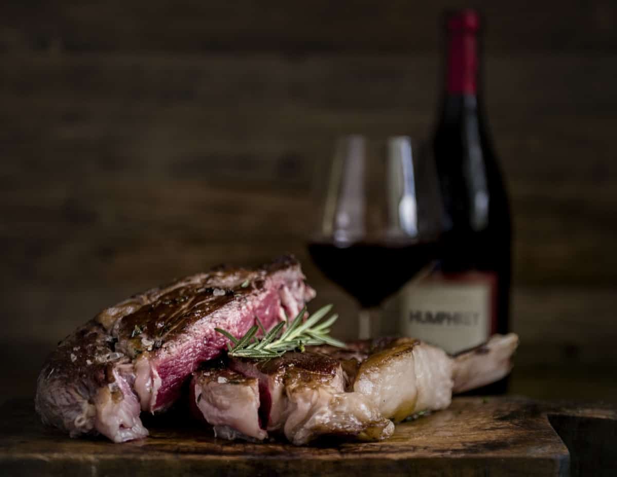 סטייק פרוס, במידת עשייה בינונית-נדירה, מעוטר ברוזמרין, מונח על קרש עץ, עם כוס יין אדום ובקבוק יין ברקע המטושטש.