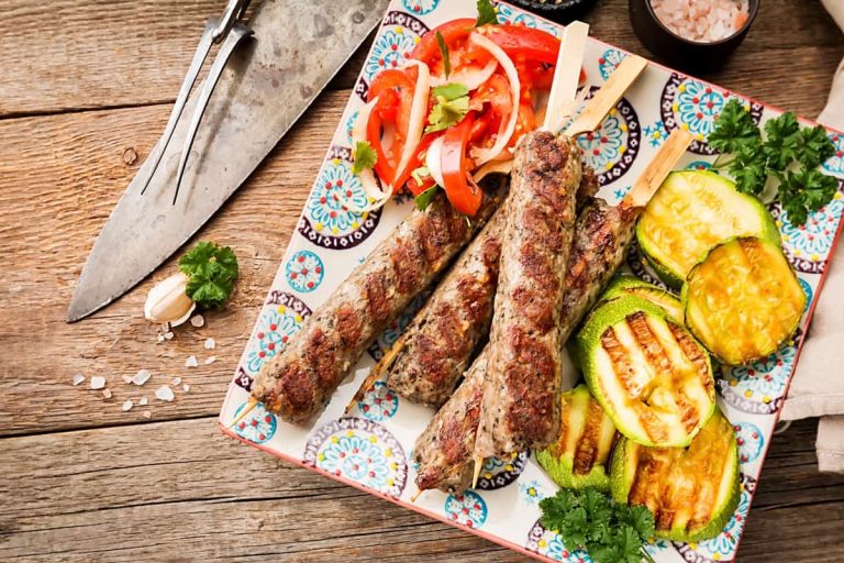 שלושה קבבים של בשר בגריל על שיפודים, מוגשים עם קישואים בגריל, סלט עגבניות פרוסות ועשבי תיבול טריים על צלחת צבעונית, המונחים על שולחן עץ כפרי עם סכו"ם ומלח בסמוך.